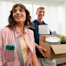 A smiling couple is moving, with the man carrying a box of household items.