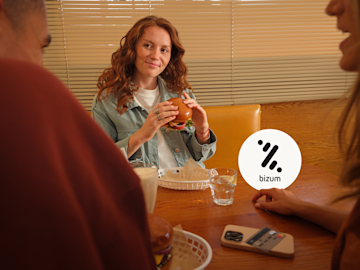 Una mujer sonríe sentada en la mesa de un restaurante con amigos mientras sostiene una hamburguesa, con el logotipo de Bizum superpuesto en la imagen.