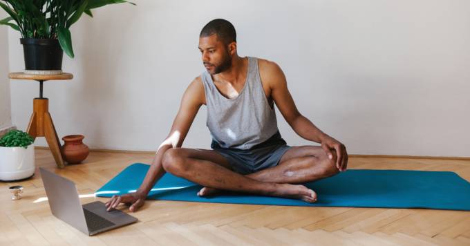 Persona sentada sobre una manta de yoga preparándose para hacer ejercicio y buscando un entrenamiento en su computadora.