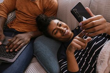 Woman lying on a sofa using her mobile and a man using his laptop.
