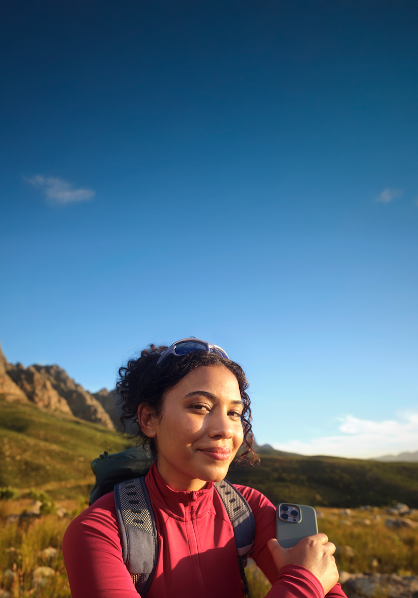 A smiling woman with a backpack holds a smartphone while standing in a mountainous landscape during golden hour.