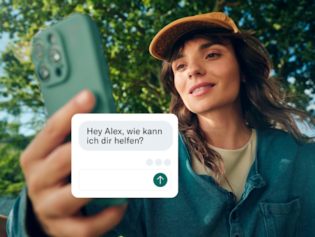 Eine Frau hält ihr Telefon in Richtung Kamera, wobei ein Kundenservice-Chatfenster über den Bildschirm gelegt ist.
