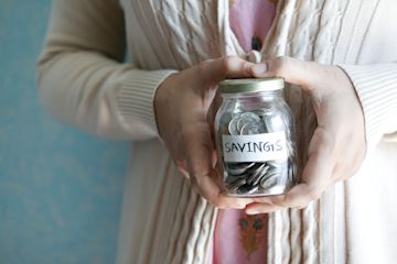 Eine Frau hält ein Glas, auf dem Ersparnisse steht, gefüllt mit Münzen.
