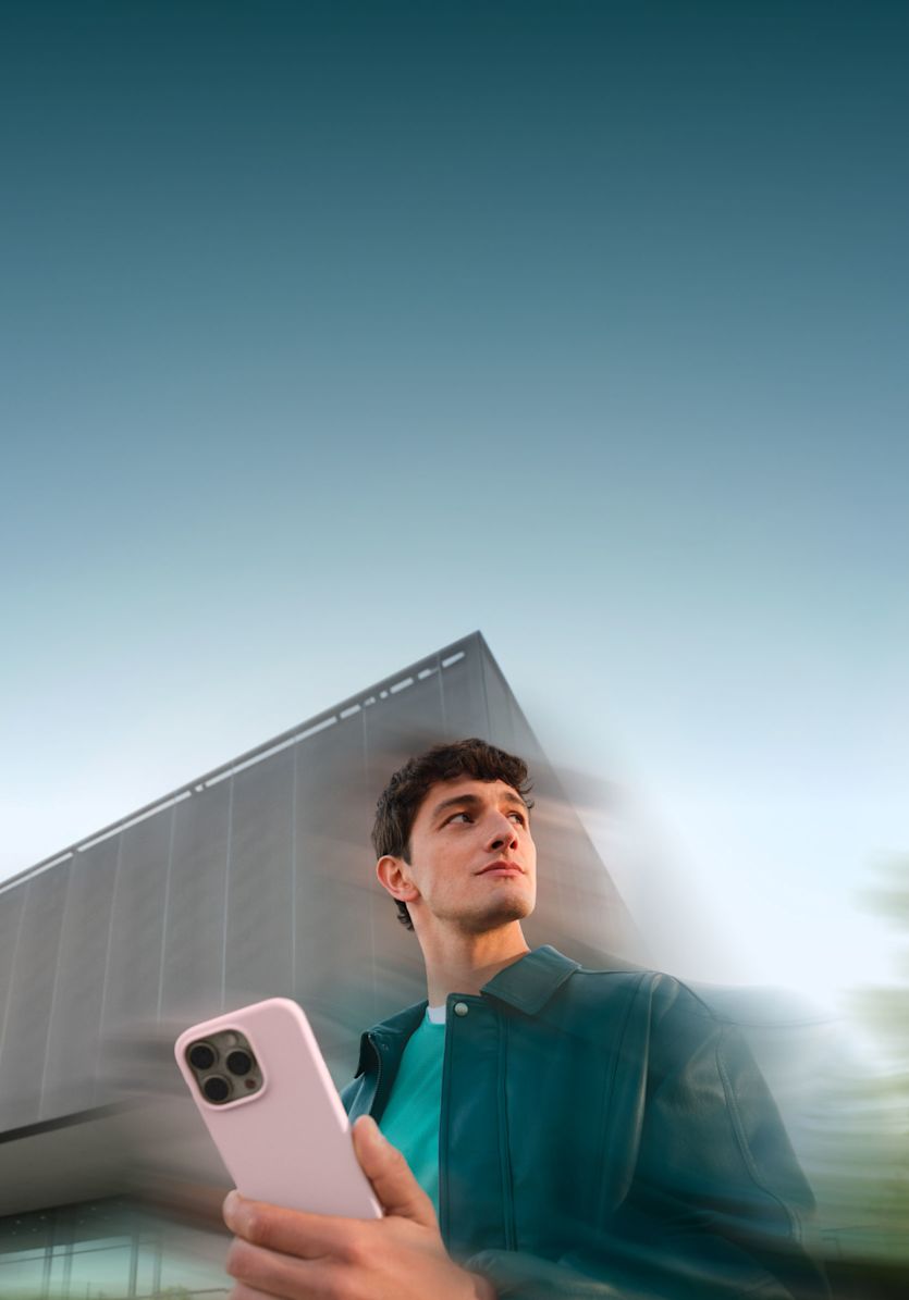 Ein Mann in einer blauen Jacke hält ein Smartphone mit einer hellrosa Hülle in der Hand und blickt nach oben und zur Seite.