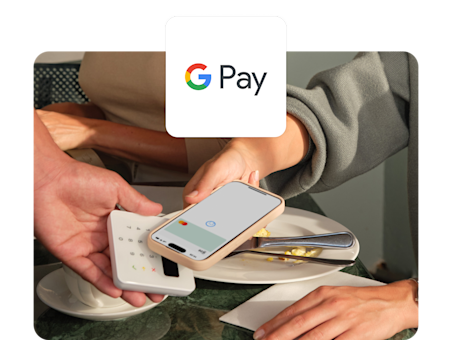 Une personne utilise un smartphone dans une coque beige pour payer à une table de restaurant via un lecteur de carte mobile avec Google Pay.