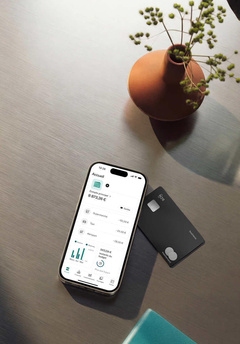 Un smartphone affichant une application bancaire est posé sur une surface en métal brossé à côté d'une carte Mastercard N26 Business noire et d'un petit vase avec des branches décoratives.