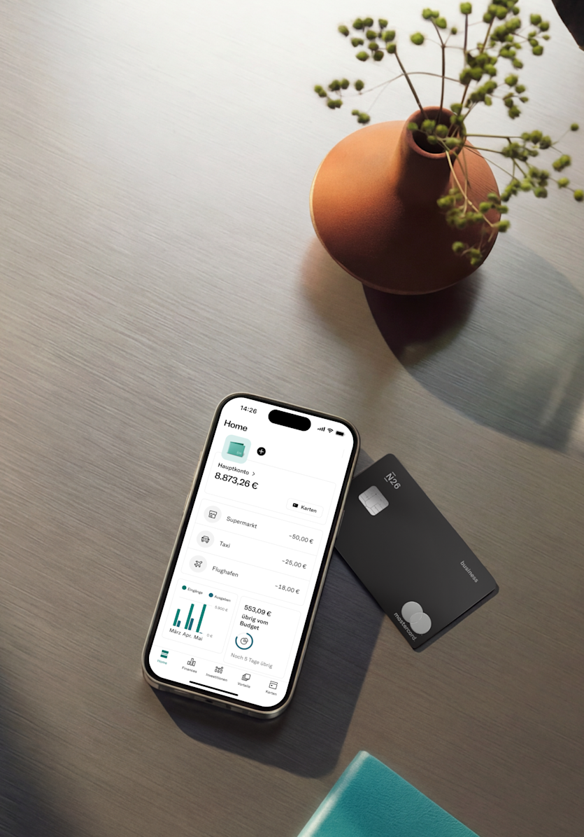 Ein Smartphone mit einer geöffneten Banking-App liegt auf einer Oberfläche aus gebürstetem Metall neben einer schwarzen N26 Business Mastercard und einer kleinen Vase mit dekorativen Zweigen.