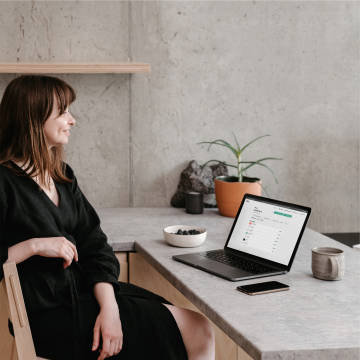 Mujer sonriente sentada frente a una mesa con un teléfono, una computadora portátil y plantas.