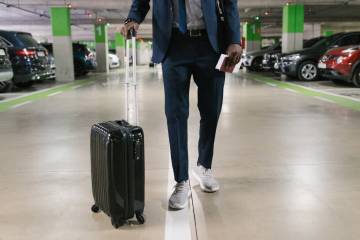 jeune homme habillé décontracté avec une valise à roulettes dans un parking à l'aéroport.