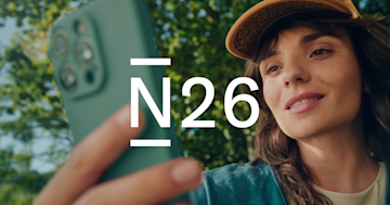 A smiling woman wearing a baseball cap holds up her smartphone outdoors.