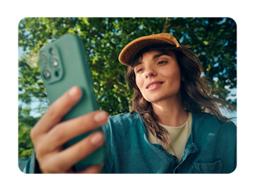 A smiling woman wearing a baseball cap holds up her smartphone outdoors.