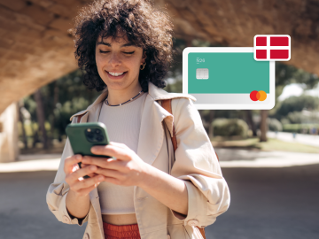 image of a young woman standing up in a park and looking at his phne, with an n26 card and a Danish flag in the background.