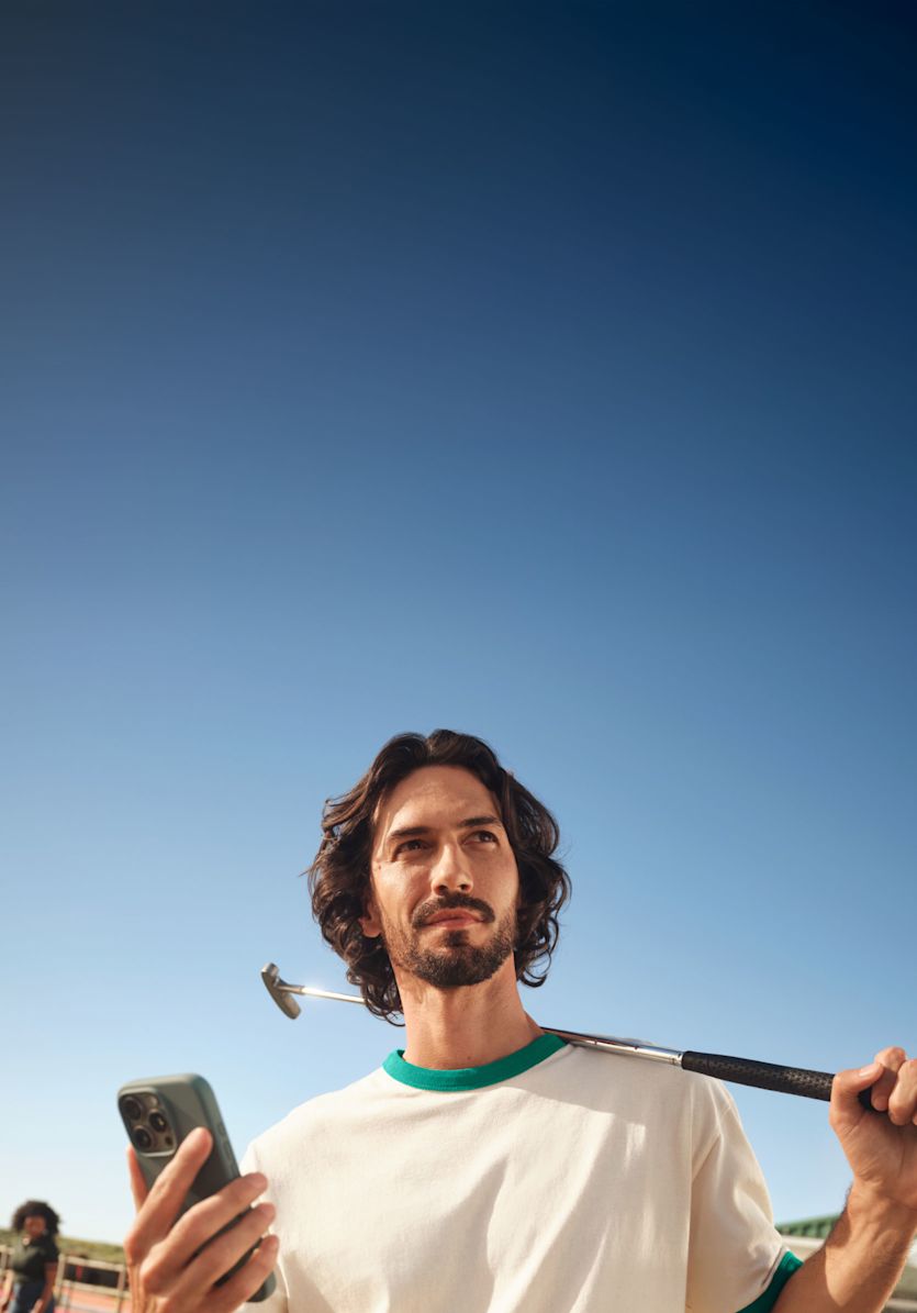 Ein Mann mit dunklen, gewellten Haaren und einem Bart steht auf einem Minigolfplatz und blickt nachdenklich in die Ferne. In der einen Hand hält er ein Smartphone, während ein Putter auf seiner Schulter ruht. Im Hintergrund sind vor einem klaren blauen Himmel andere Personen zu sehen, die in der Ferne unscharf Minigolf spielen.