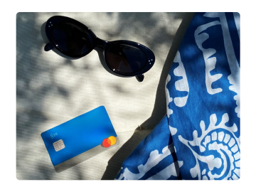 Tarjeta de color gasolina N26 y gafas de sol junto a una manta de playa en la arena.
