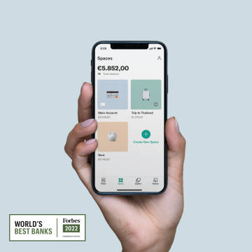 A hand holding an iPhone X in their hand with the Spaces screen open on the N26 app with Forbes best bank logo.