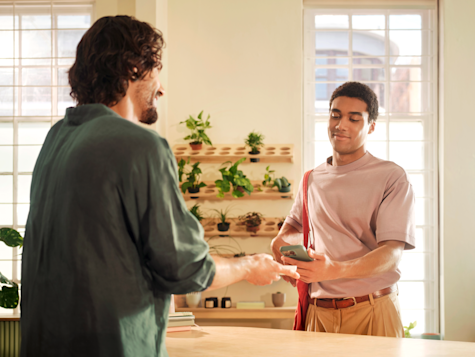 A man behind a counter hands a card to a customer who is looking at his smartphone.