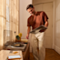 Un homme en t-shirt marron sourit en tenant un disque vinyle à côté d'une platine dans une pièce ensoleillée.