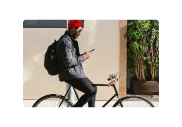 Un caballero con un gorro rojo y una chaqueta de mezclilla, sentado en una elegante bicicleta negra, revisa su teléfono. Su mochila está lista para el viaje, y está a solo unos pasos de un edificio con una encantadora planta en maceta.