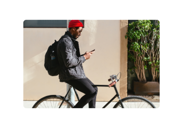 Un gentiluomo con un berretto rosso e una giacca di jeans, appollaiato su una bicicletta nera elegante, controlla il suo telefono. Il suo zaino è pronto per il viaggio, ed è a pochi passi da un edificio con una pianta in vaso incantevole.
