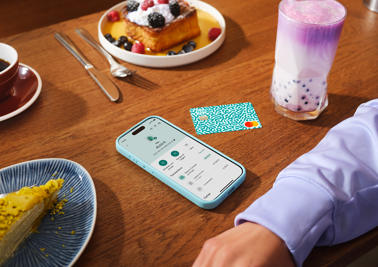 Un téléphone portable affichant l'interface « Moins de 18 ans » de l'application N26, à côté d'une carte de débit physique posée sur une table en bois avec un petit-déjeuner.