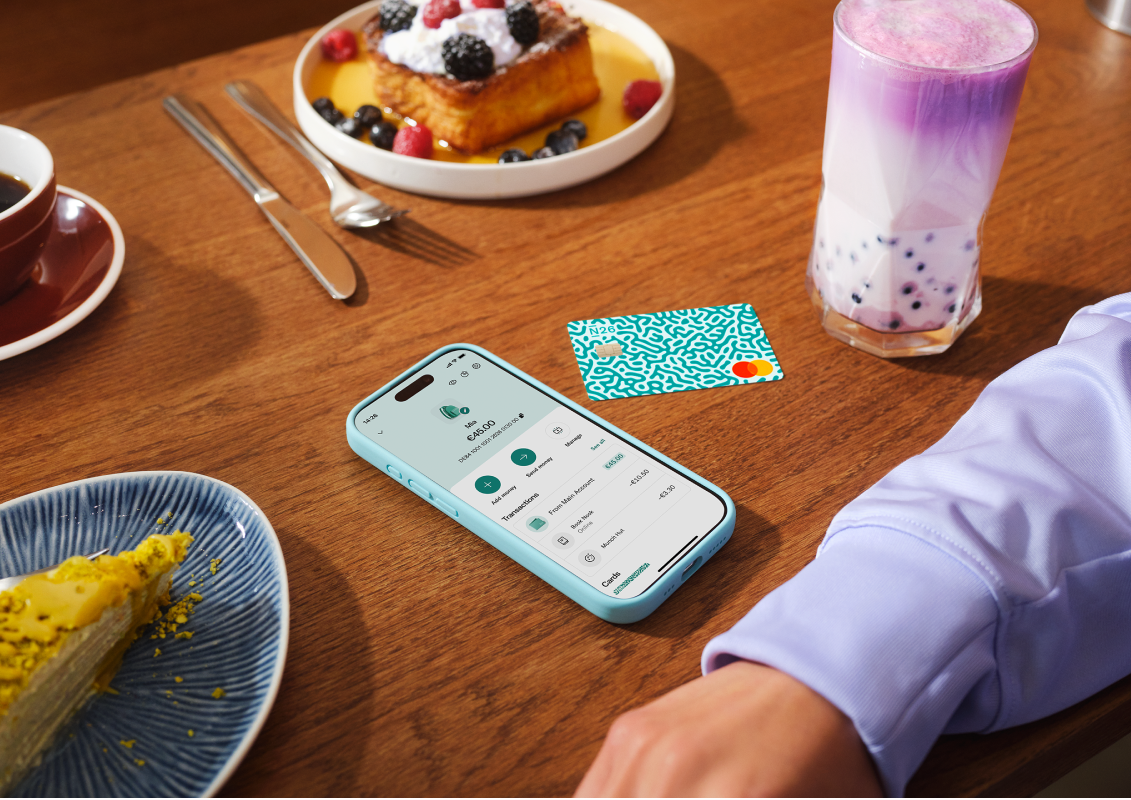 A mobile phone showing the N26 app "Under 18" interface next to a physical debit card on a wooden table with breakfast.