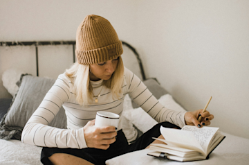 A woman dressed in winter clothes writing on her bed. 