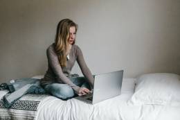 Frau sitzt auf einem Bett und benutzt ihren Laptop.