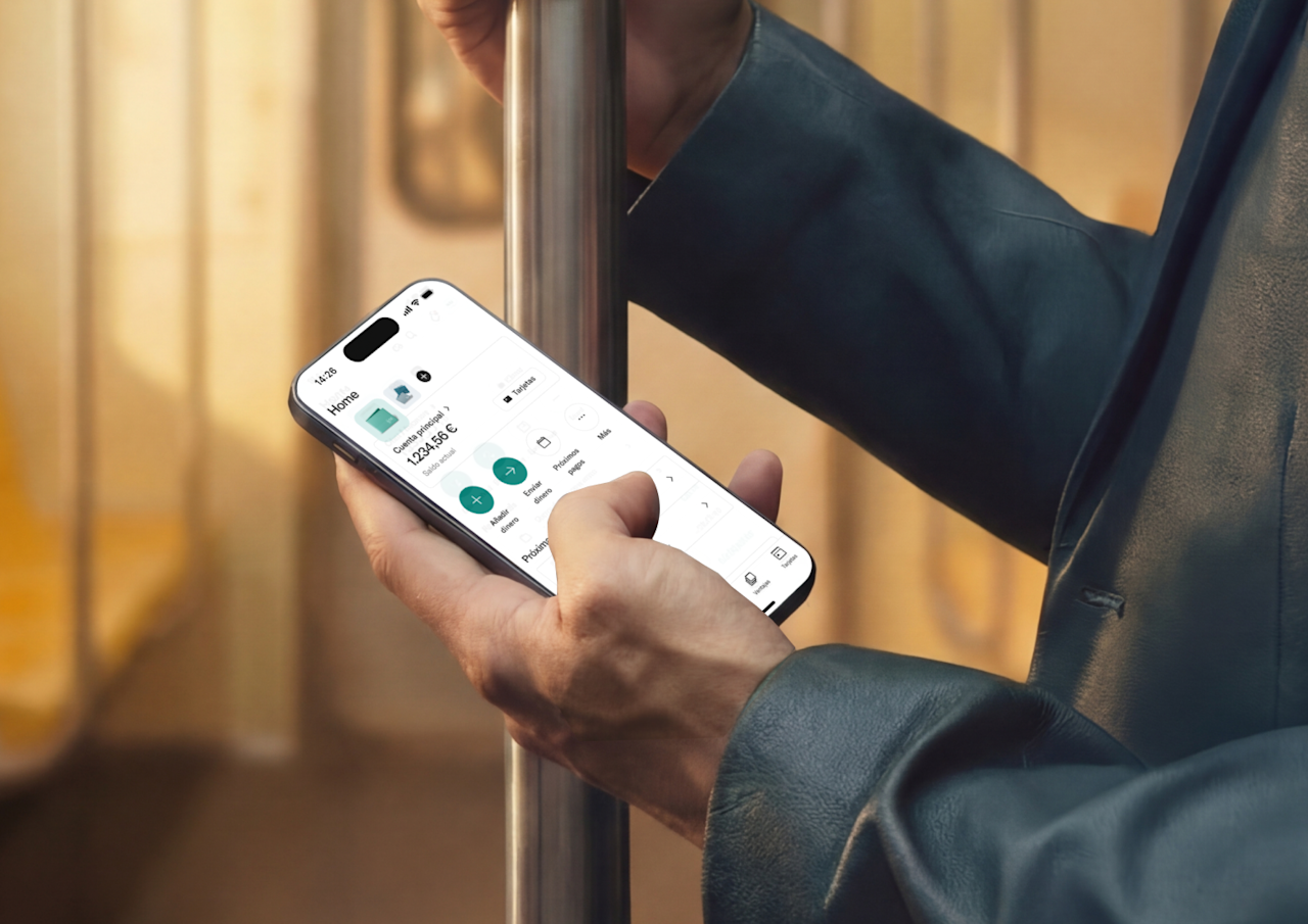 Una persona se sujeta a una barra de metal en un tren mientras utiliza un teléfono inteligente che muestra la interfaz de una aplicación de banca móvil.