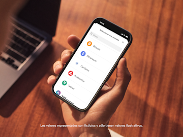 Una mano sostiene un smartphone que muestra la pantalla de selección de criptomonedas para comprar una moneda.