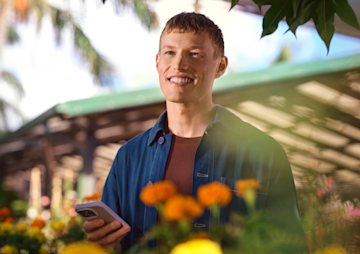 Un hombre sonriente sostiene un smartphone mientras está de pie al aire libre rodeado de flores amarillas y naranjas.