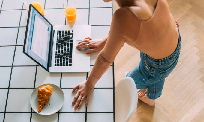 Woman stoops over laptop standing between an orange juice and a croissant.