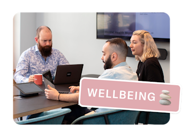 Three people are gathered around a table with laptops and tablets, seemingly in a meeting, with the word "WELLBEING" and a stack of stones overlaid on the image.