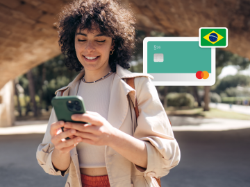 image of a young woman standing up in a park and looking at his phne, with an n26 card and a Brazilian flag in the background.