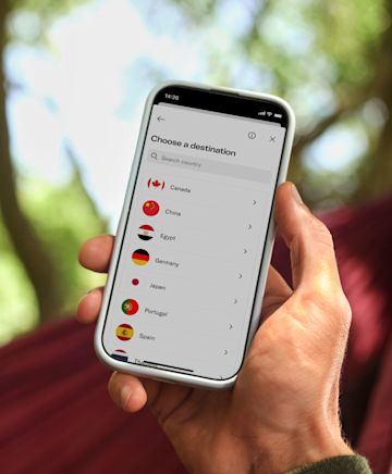 A person is holding a smartphone outdoors, with green trees blurred in the background. On the phone screen, an app is open showing a menu titled “Choose a destination”, with a search bar and a list of countries displayed. Visible options include Canada, China, Egypt, Germany, Japan, Portugal, and Spain, each accompanied by its national flag. The scene suggests someone planning or preparing for international travel.