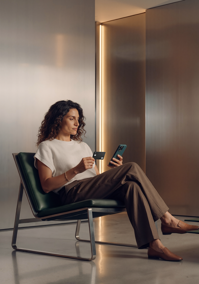 Une femme est assise dans una chaise vert foncé dans une pièce minimaliste, tenant une carte N26 Metal noire tout en regardant son smartphone.