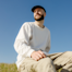 A man wearing a white sweatshirt and a black cap is sitting outdoors in a sunny field.