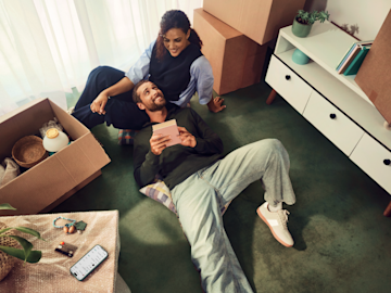 A couple is sitting on the floor surrounded by moving boxes, looking at a small pink notebook together.