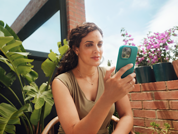 Una mujer se sienta en un balcón soleado y mira su smartphone con una funda de color verde azulado.