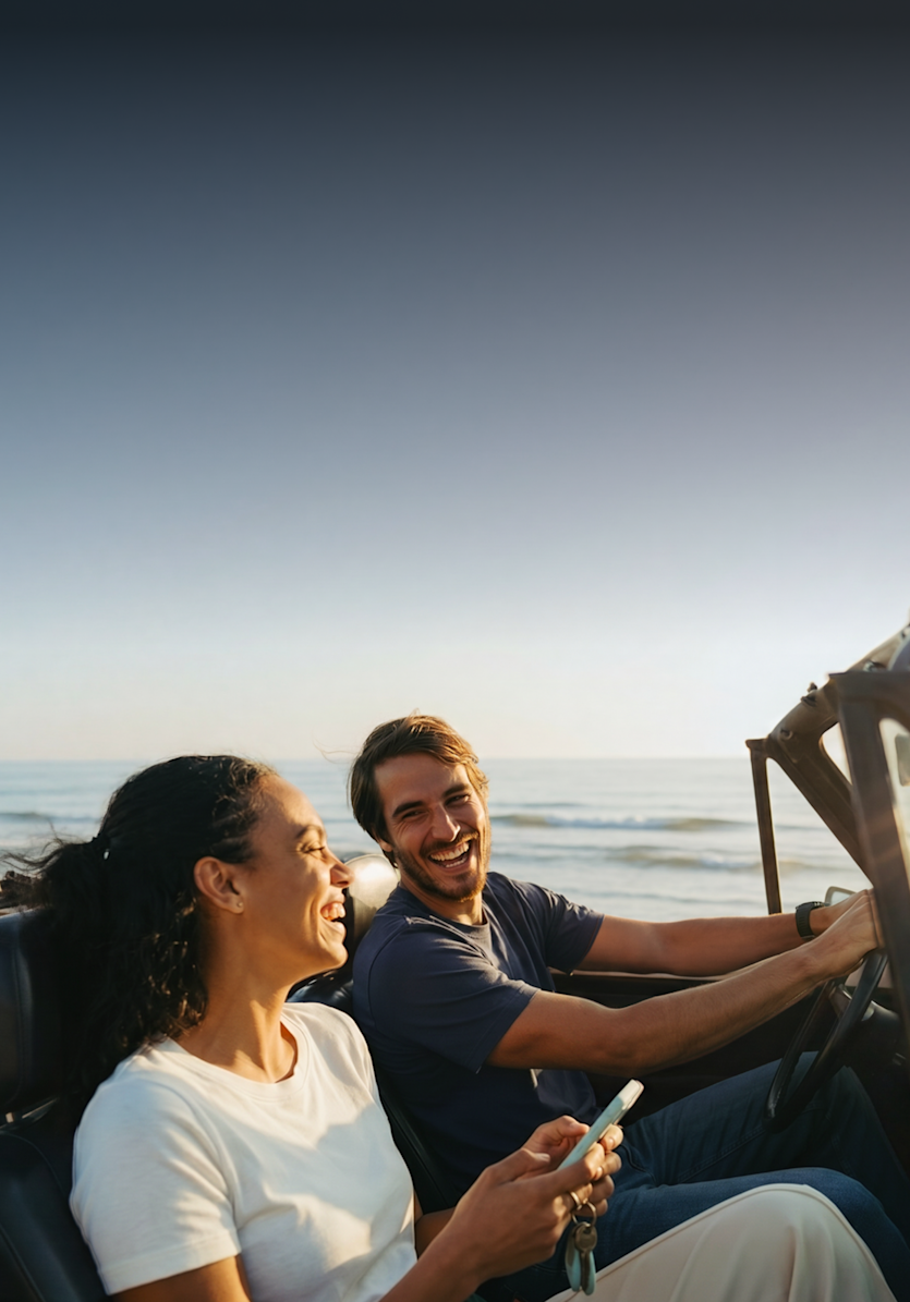 Ein lächelnder Mann und eine Frau fahren an einem sonnigen Tag in einem Cabrio entlang einer Strandküste.