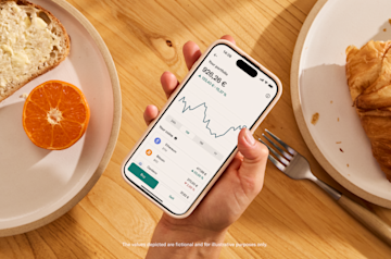 A hand holds a smartphone displaying a cryptocurrency portfolio with a line graph and balances at a table with breakfast food.