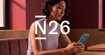 Una mujer sentada en el banco de un restaurante mira su teléfono inteligente, con un gran logotipo blanco de N26 superpuesto en primer plano.