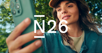 Una mujer con sombrero mira un teléfono inteligente en su mano al aire libre con el logotipo de N26 superpuesto en el centro.