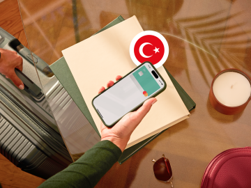 image of a person holding a phone showing an N26 card in the app screen and grabbing a suitcase with the other hand and with a turkish flag.