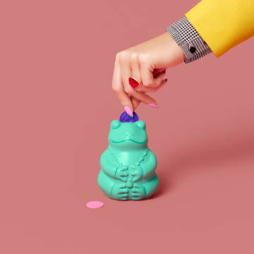 A hand putting plastic coins into a mint colored frog piggy bank.