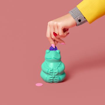 A hand putting plastic coins into a mint colored frog piggy bank.