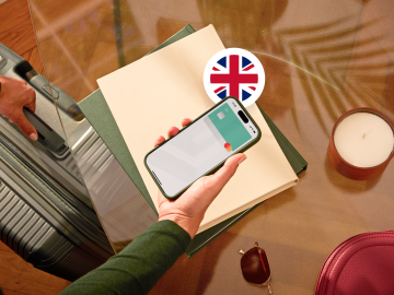 image of a person holding a phone showing an N26 card in the app screen and grabbing a suitcase with the other hand and with a British flag.