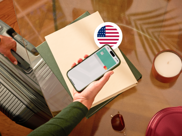 image of a person holding a phone showing an N26 card in the app screen and grabbing a suitcase with the other hand and with a USA flag.