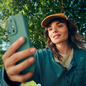 Una mujer sonriente con gorra de béisbol sostiene su smartphone en alto al aire libre.