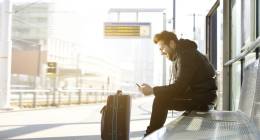giovane uomo seduto in una stazione ferroviaria con un bagaglio e controllando il suo telefono cellulare.