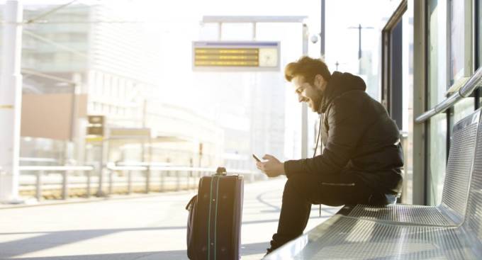 junger Mann mit einem Koffer in einem Bahnhof sitzt und mit seinem Handy überprüft.